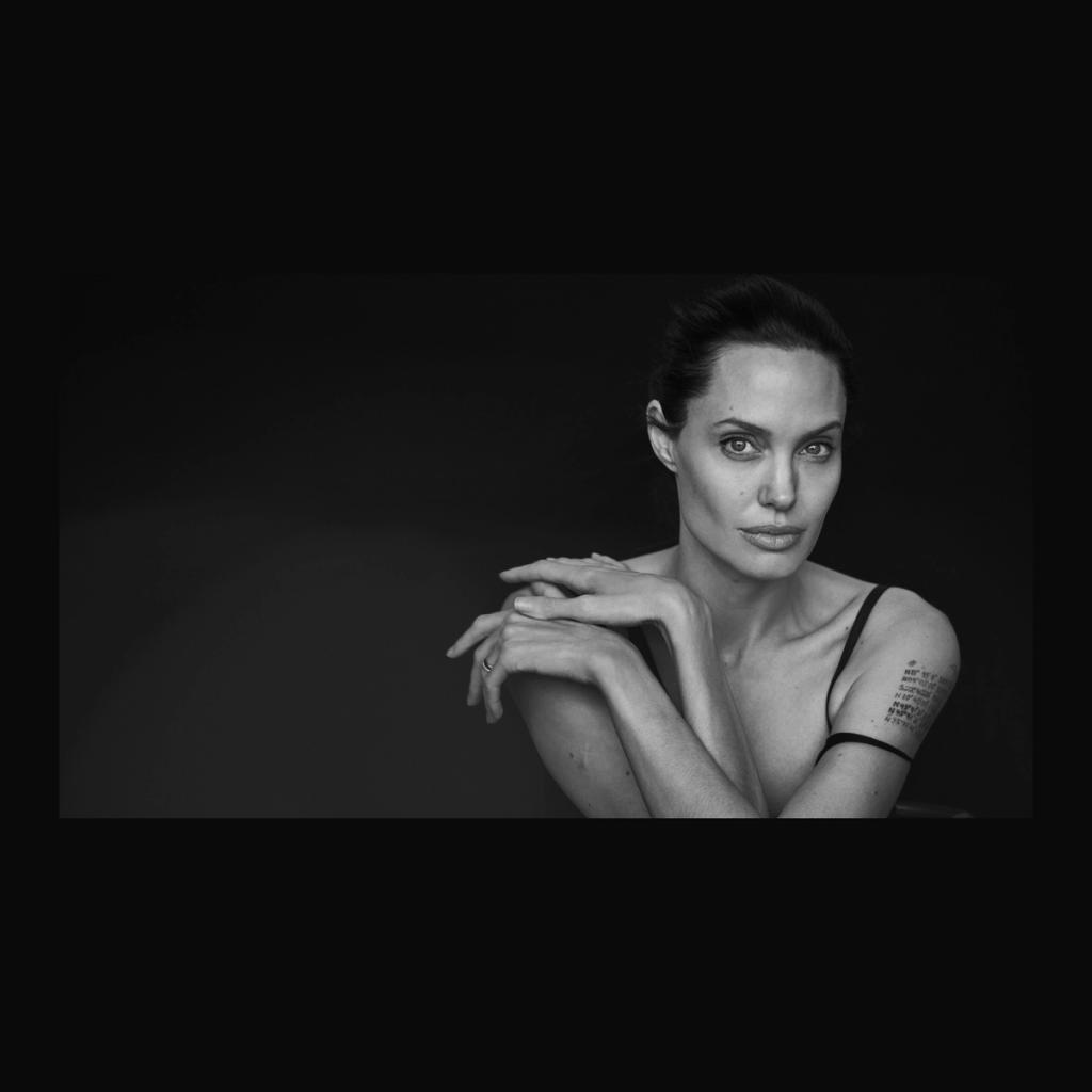 A black-and-white portrait of a woman resting her chin on her folded hands, with a tattoo on her shoulder