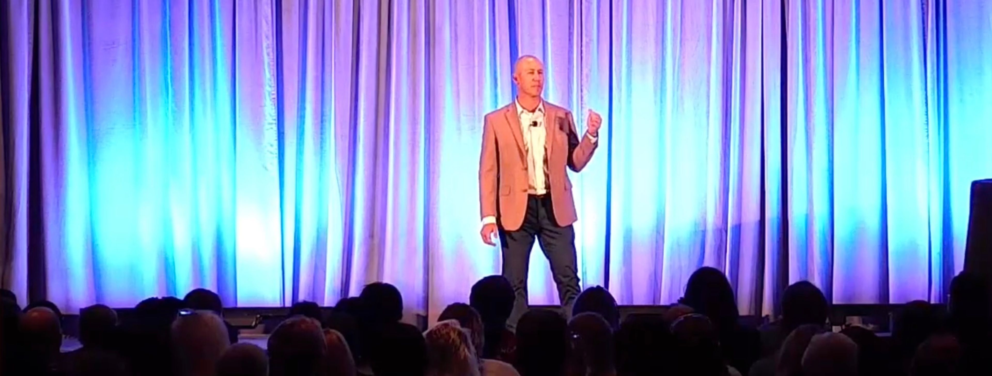 Taylor Candella on stage, captured mid-speech with a wide view of the venue lighting and audience.