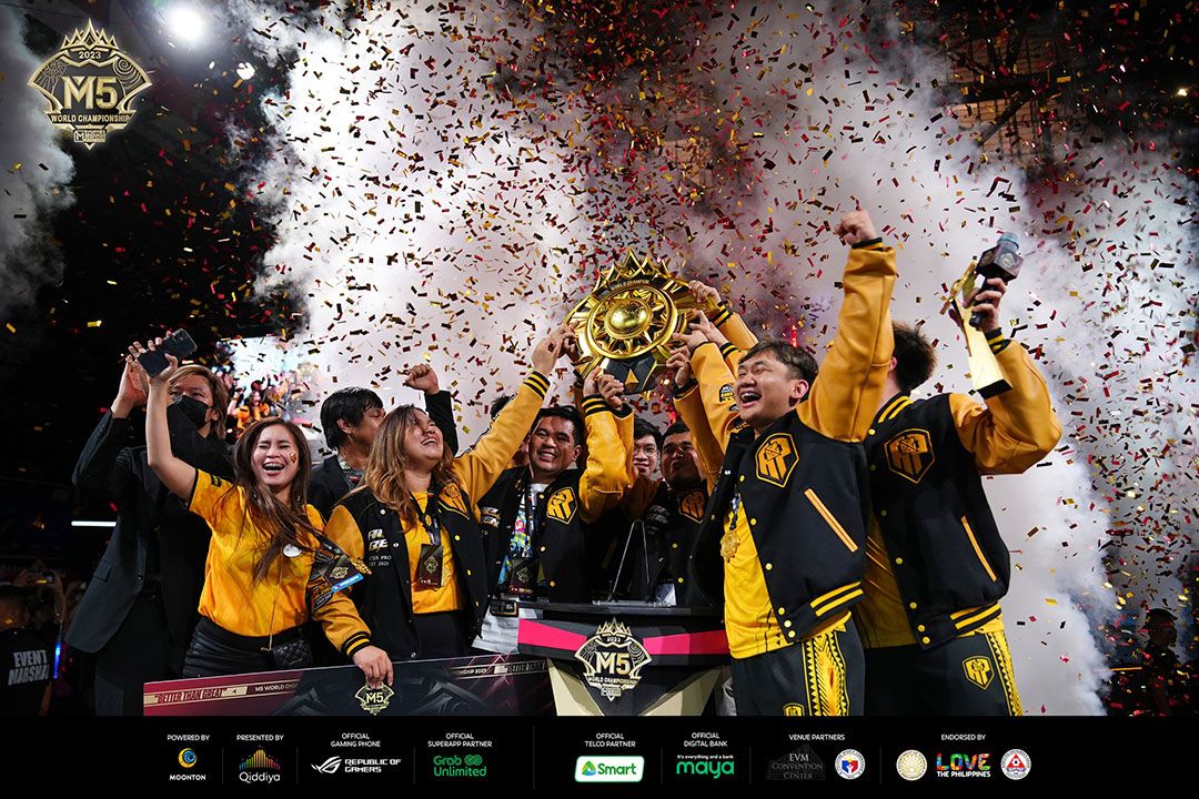 A victorious esports team AP Bren in yellow and black jackets celebrates their win with joy and excitement. They are lifting a golden trophy above their heads amidst a shower of colorful confetti. The atmosphere is festive and triumphant, capturing the climax of their success at the M5 World Championship, as indicated by the logo in the background. The stage is adorned with sponsor logos and the event is well-attended, indicating the significance of the occasion in the esports community.