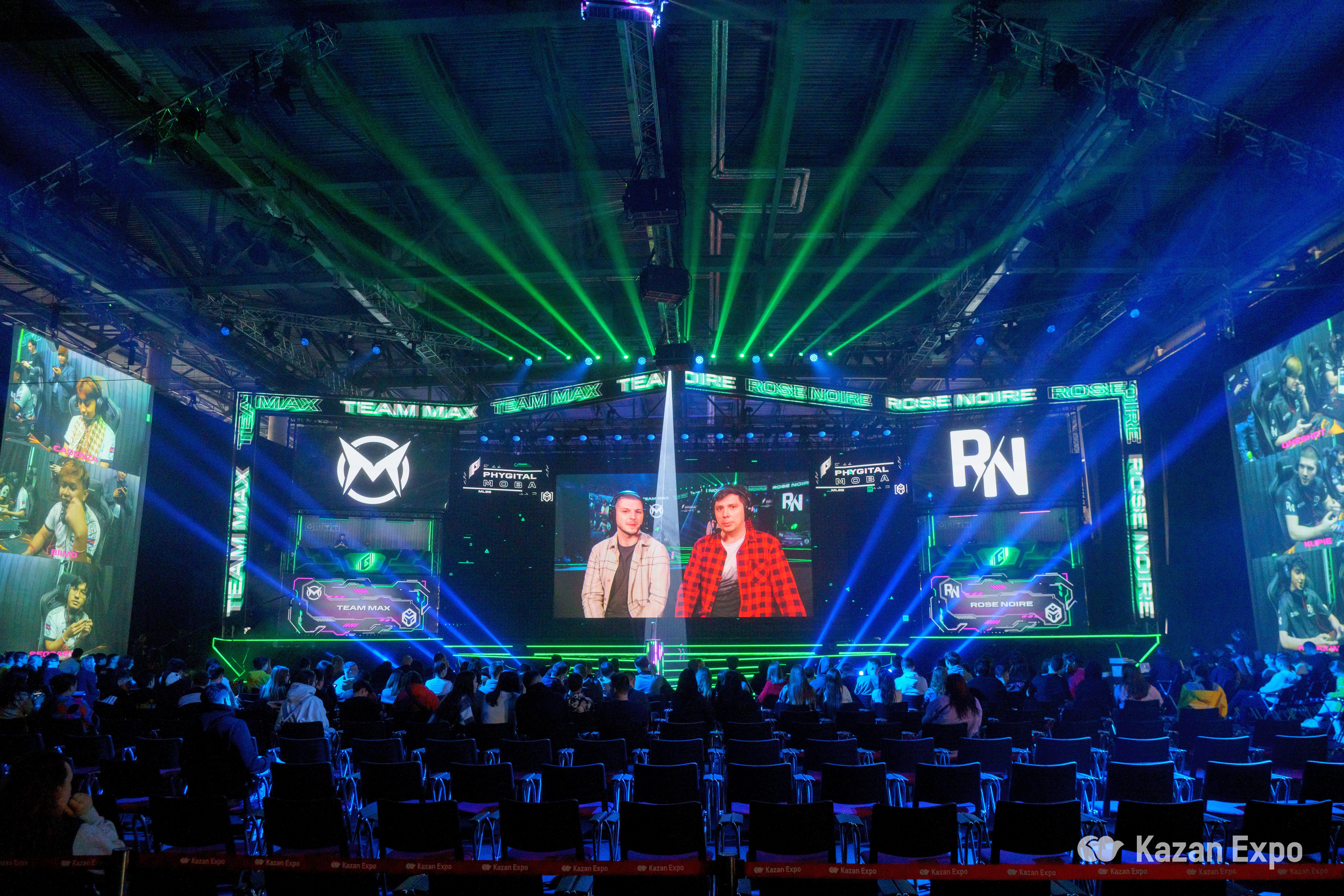 The Kazan Expo arena is illuminated with vibrant green stage lights and large screens displaying team logos, "TEAM MAX" and "ROSE NOIRE", during an esports event. The audience seats are partially filled, with attendees focused on the screens that show live gaming action and commentators discussing the ongoing match. The atmosphere is lively with the excitement of competitive gaming.