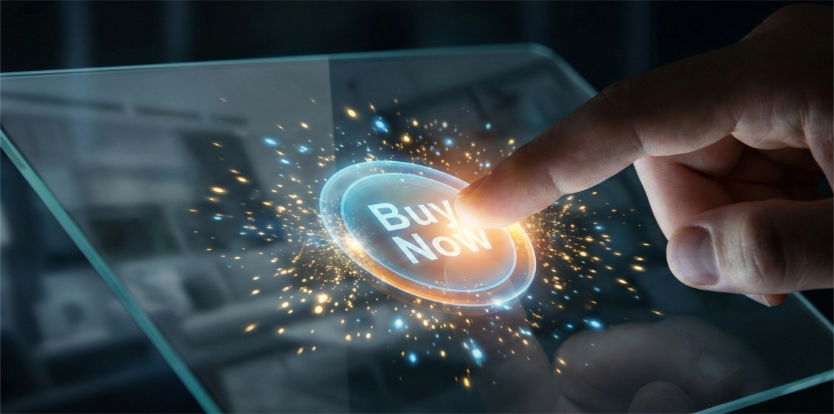 A close-up photograph of a person's index finger pressing a glowing, virtual "Buy Now" button on a transparent futuristic tablet screen. The interaction causes a burst of blue and orange digital light particles to explode from the touchpoint against a dark, blurred background.