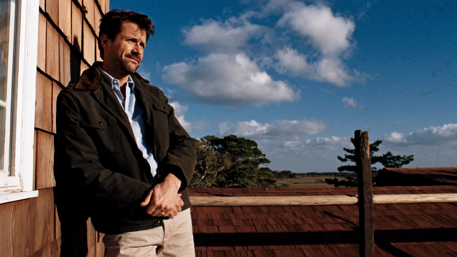Man around 40 in a waxed jacket leaning on a sunny wooden deck, illustrating private health insurance (PKV) for people over 40.