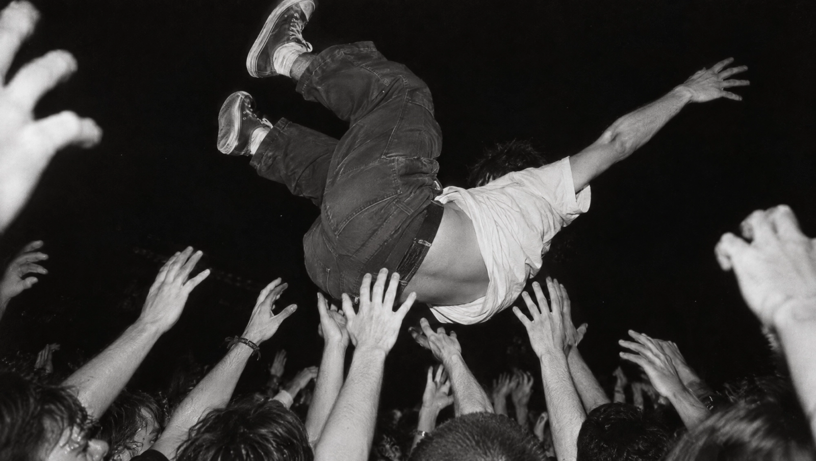 A young man crowd surfing upside down, caught by many outstretched hands, in black and white with flash
