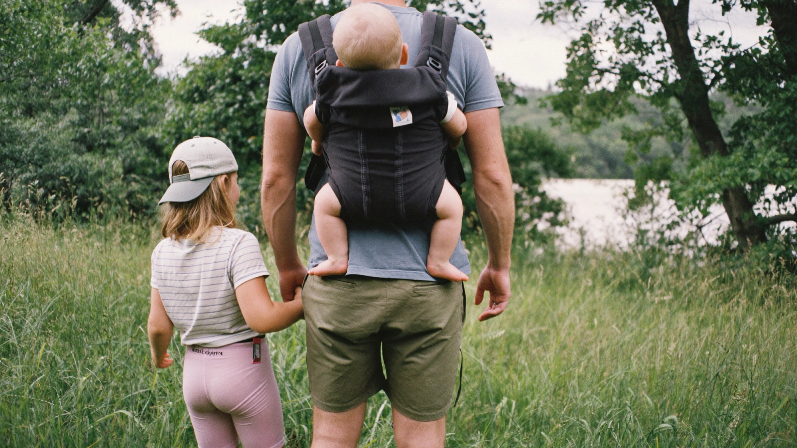A father seen from behind with a baby in a carrier on his back, holding his daughter’s hand at the edge of a green meadow.