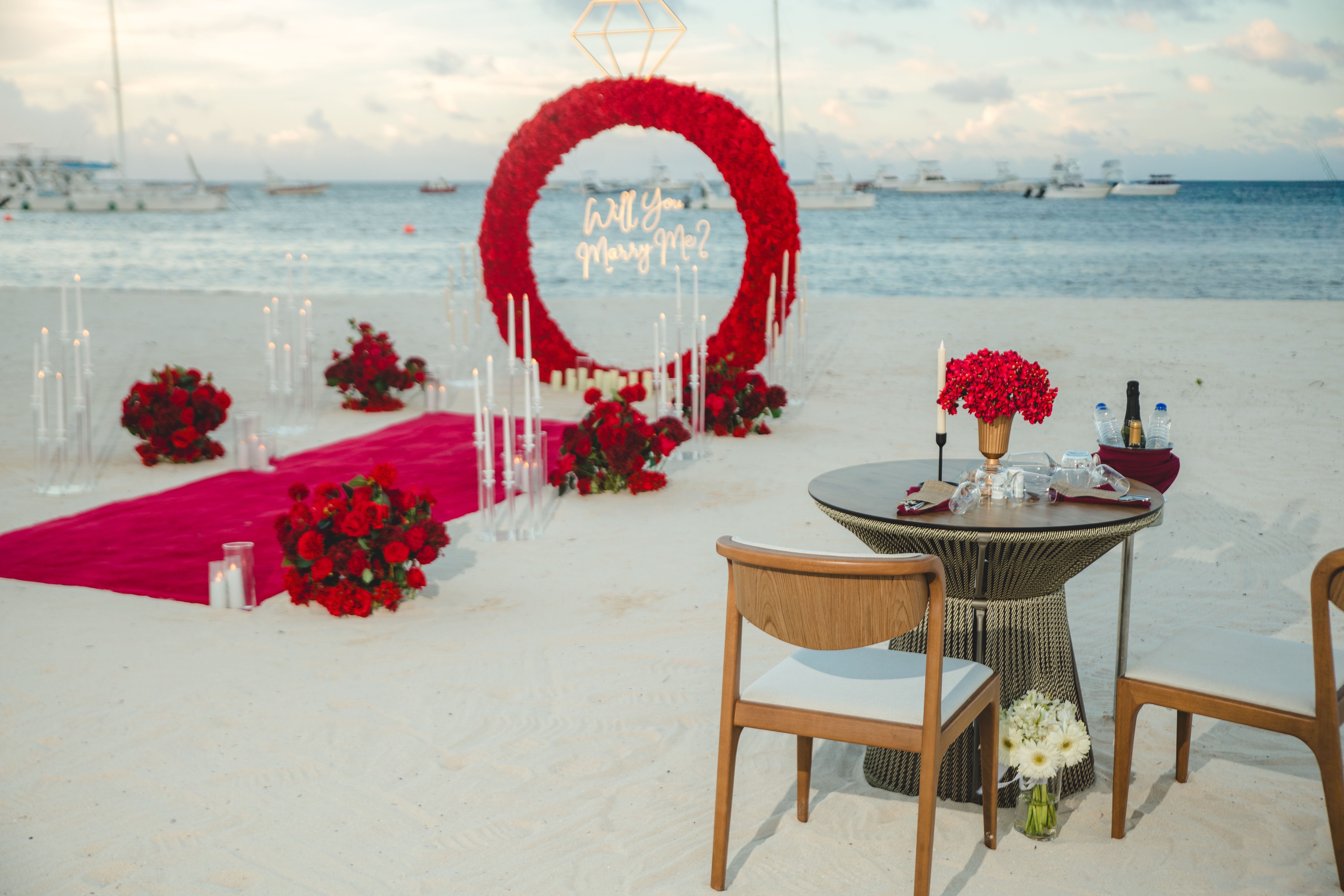 Red floral engagement ring with an illuminated diamond, proposal sign and candles by the sea
