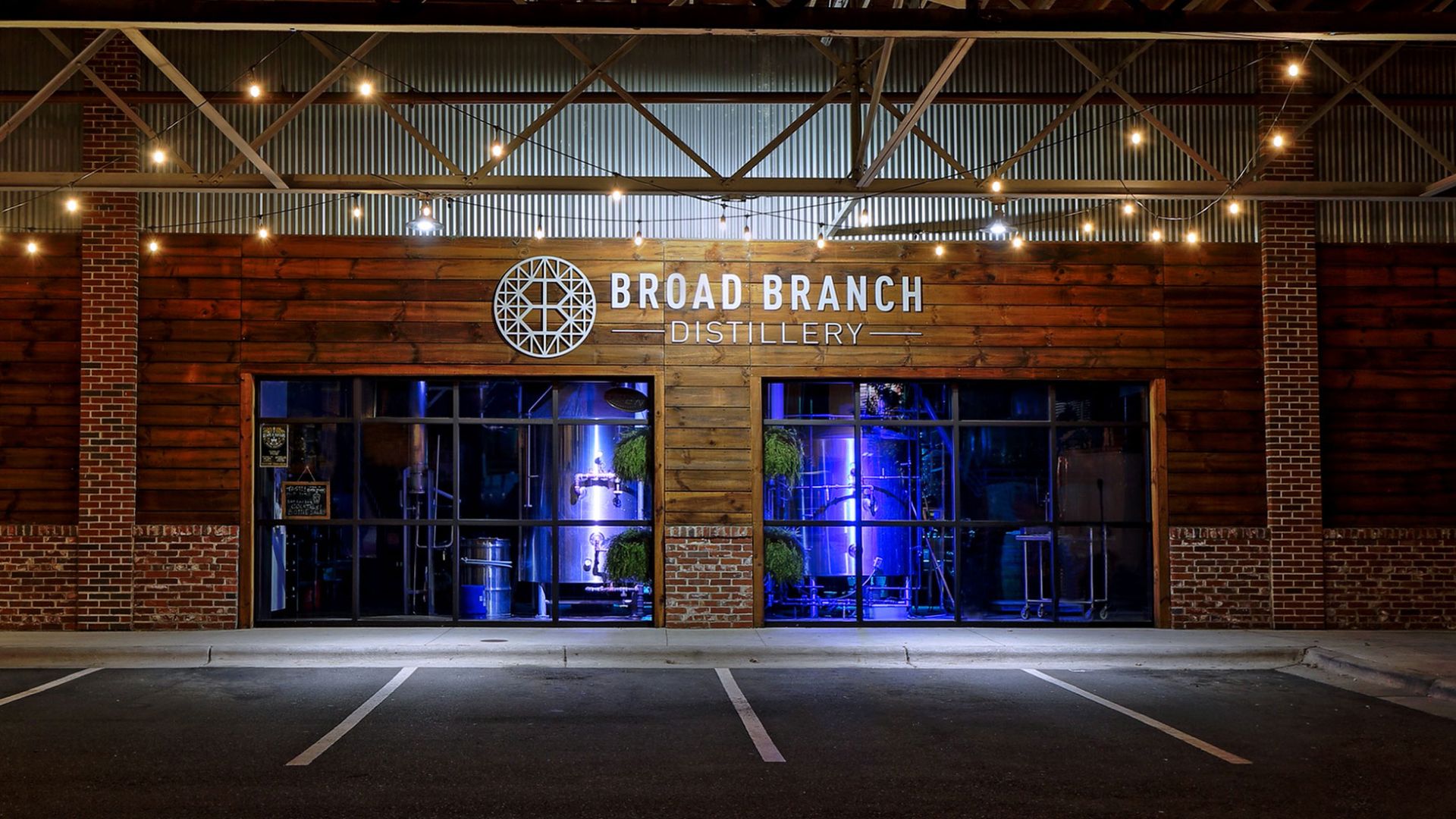 Broad Branch Distillery storefront at night with illuminated glass doors and copper distilling tanks