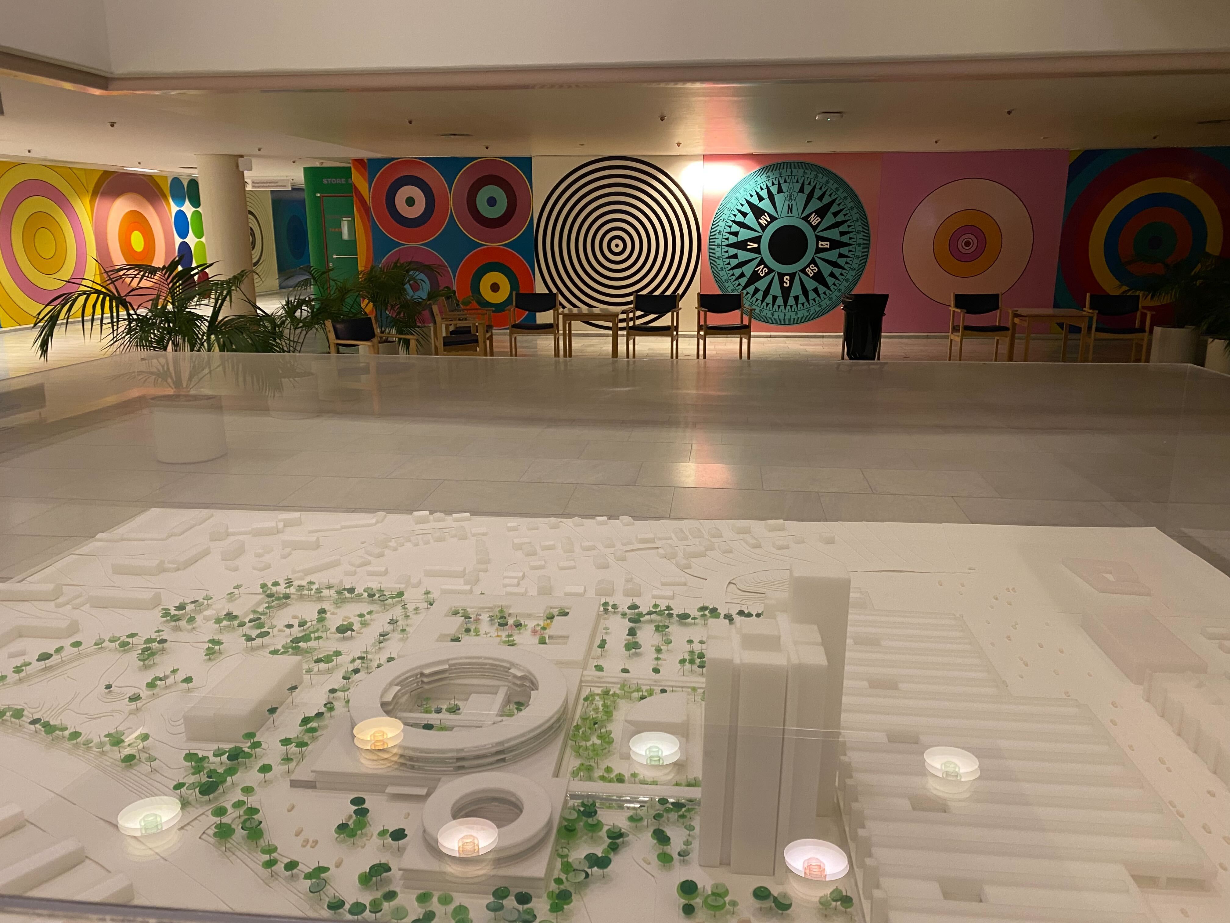 Photo from a large waiting area with a model of the Herlev Hospital complex. Walls are painted with figurative motives in bright colors by Paul Gernes