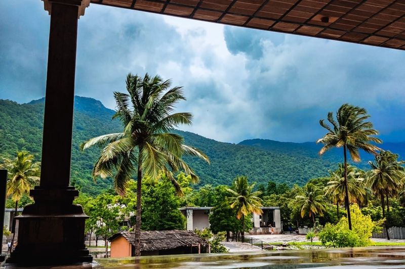 View of lush green hills and palm trees from a traditional open practice hall beneath monsoon clouds.
