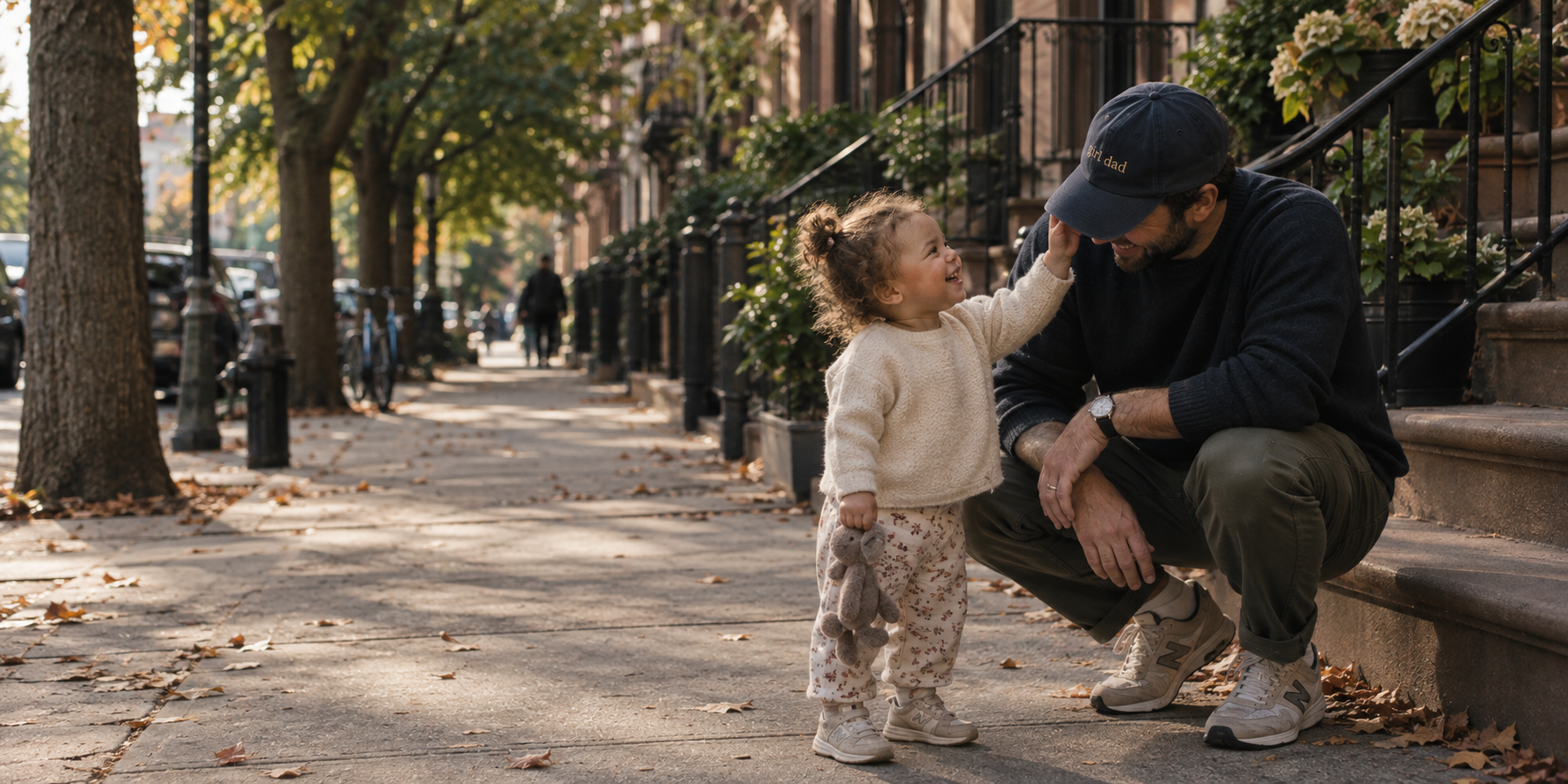 A father crouched on a sunlit Brooklyn sidewalk laughing with his young daughter