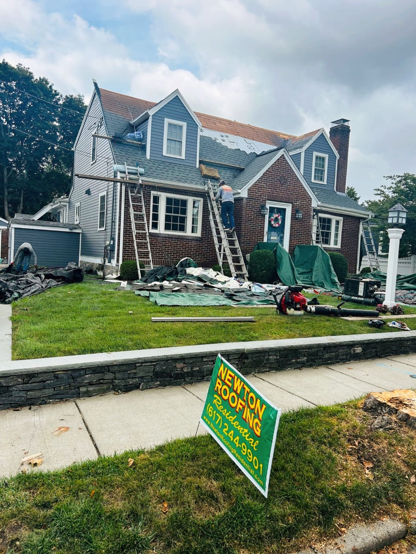 A Cape-style house mid tear-off, the front slope stripped back to bare deck with tarps laid over the shrubs and a Newton Roofing lawn sign at the curb