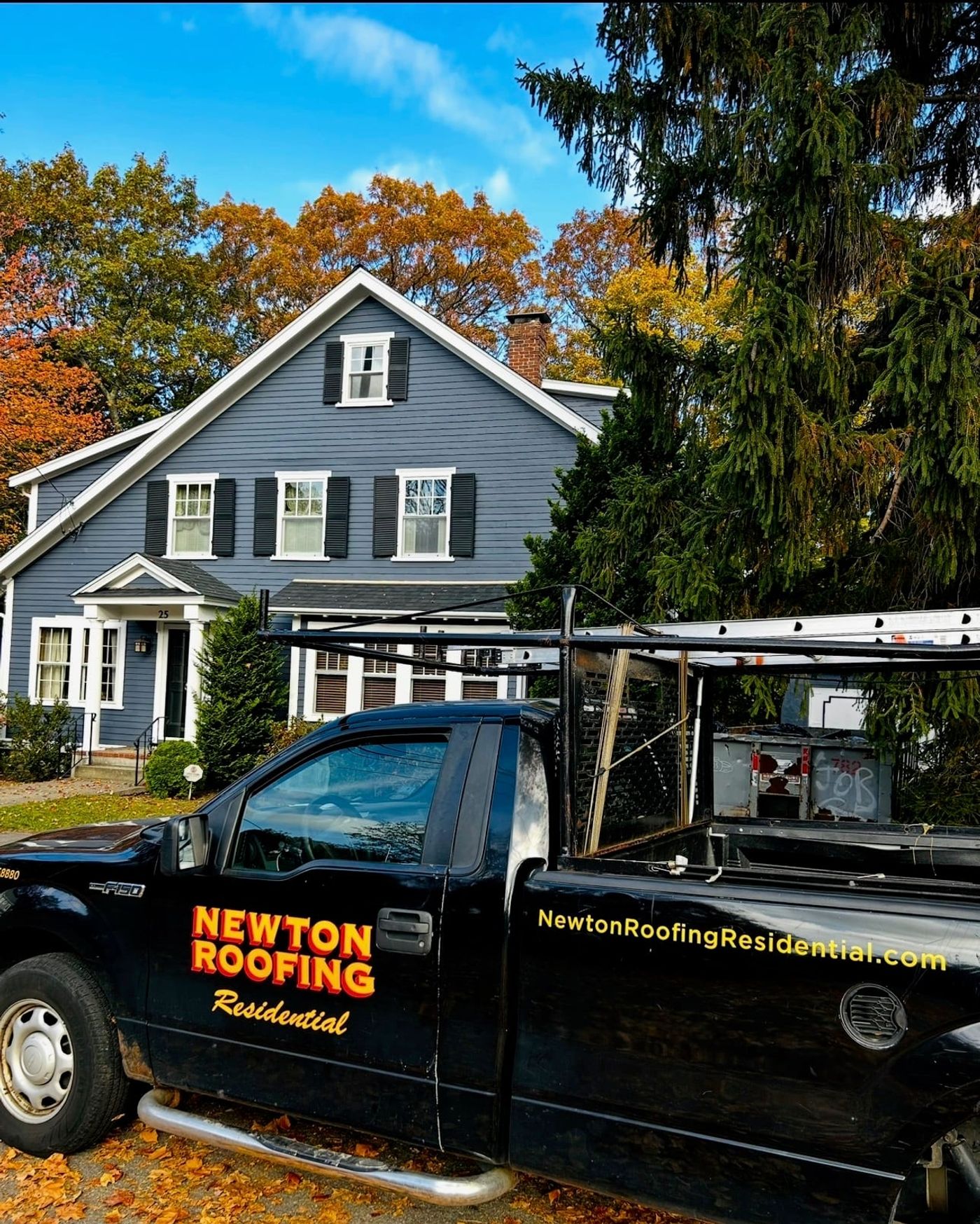 A branded Newton Roofing Residential pickup loaded with ladders parked outside a gray colonial, with fall foliage behind
