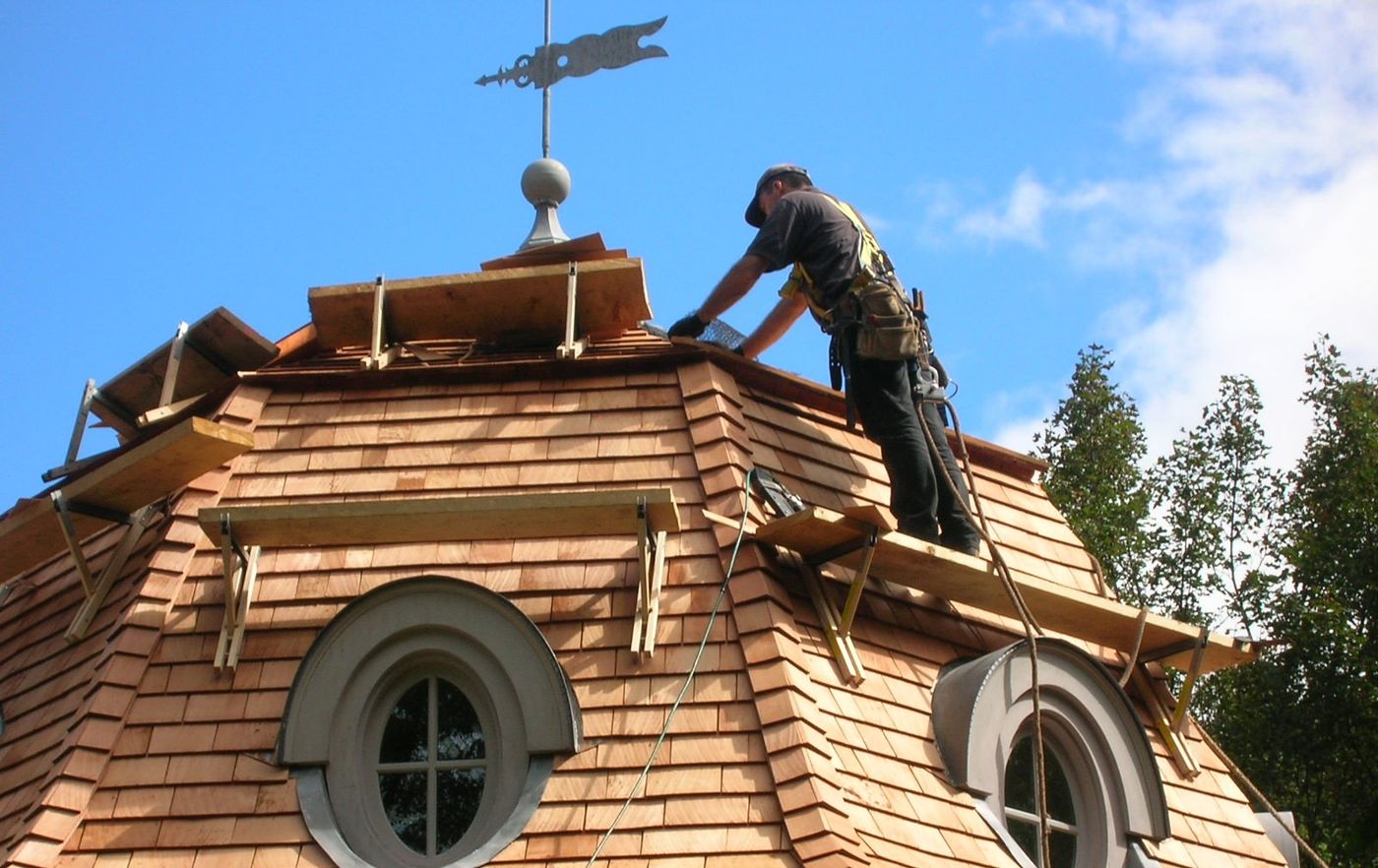 Roofer working from staging on a partly finished cedar shingle turret beneath a weather vane