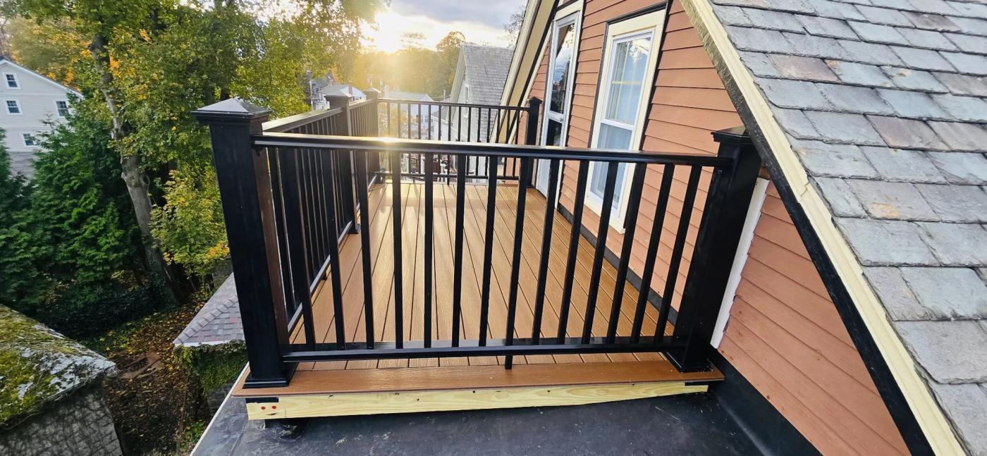 A composite rooftop deck with black aluminum railings built against a slate roof slope, lit by low evening sun