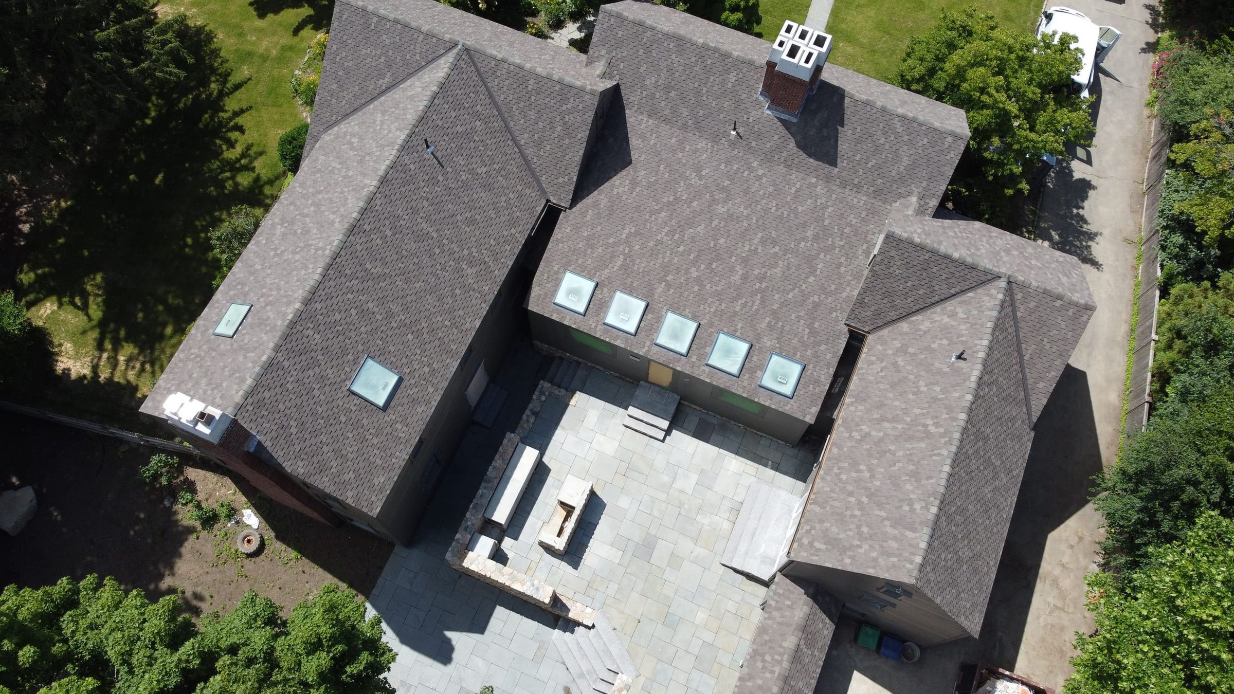 Aerial view of a completed asphalt shingle roof with five skylights and a brick chimney, over a bluestone courtyard