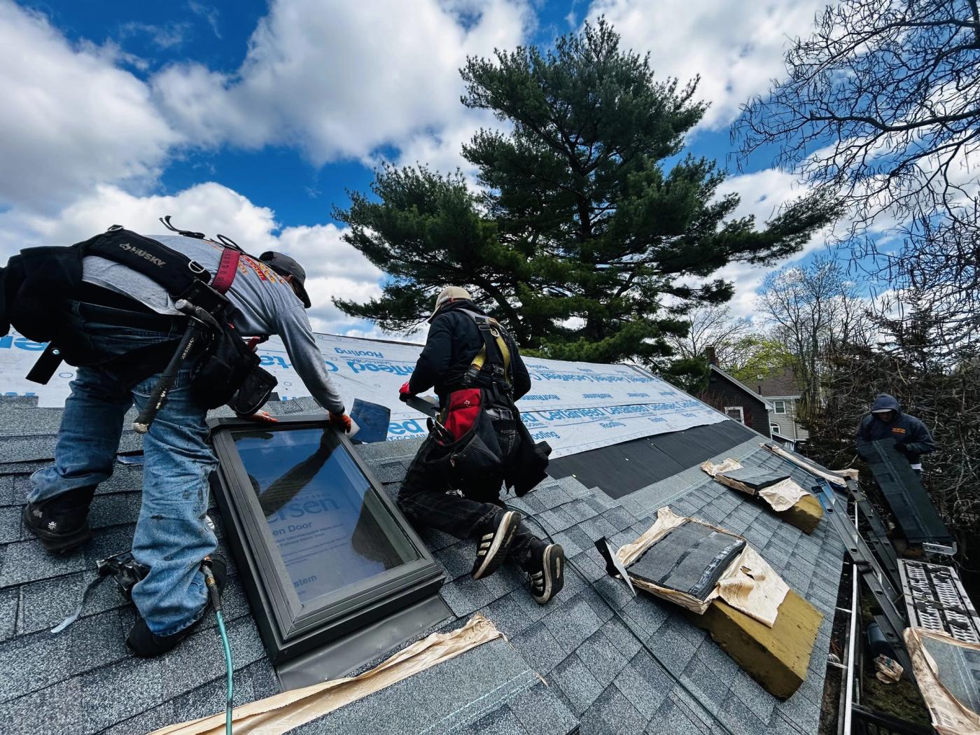 Two Newton Roofing crew members in full fall-arrest harnesses setting a new skylight into a stripped roof deck under a bright sky