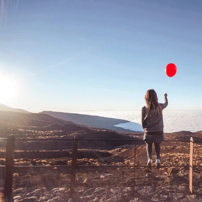 Mujer con globo rojo observa un paisaje montañoso y nubes al atardecer.