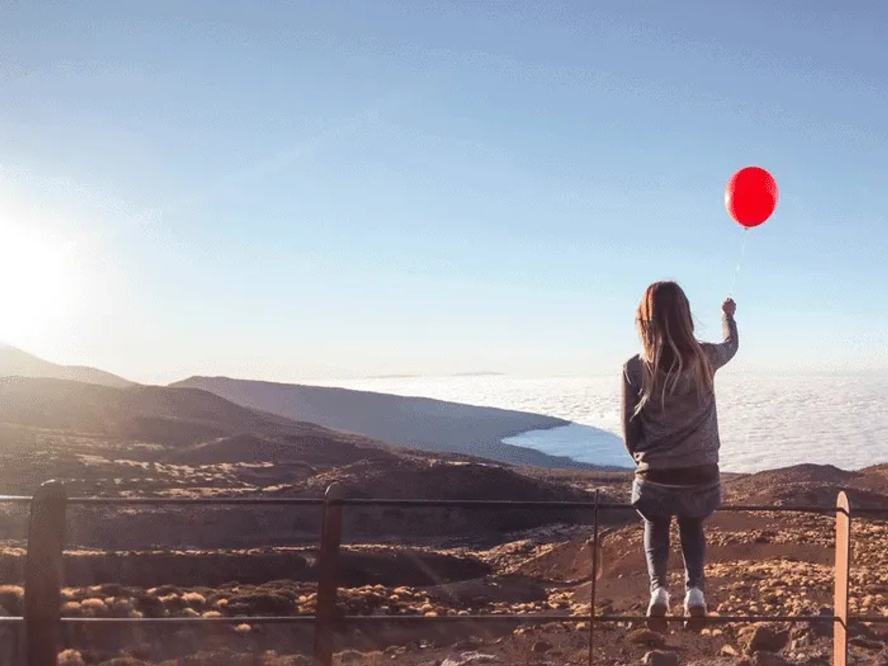 Mujer con globo rojo observa un paisaje montañoso y nubes al atardecer.