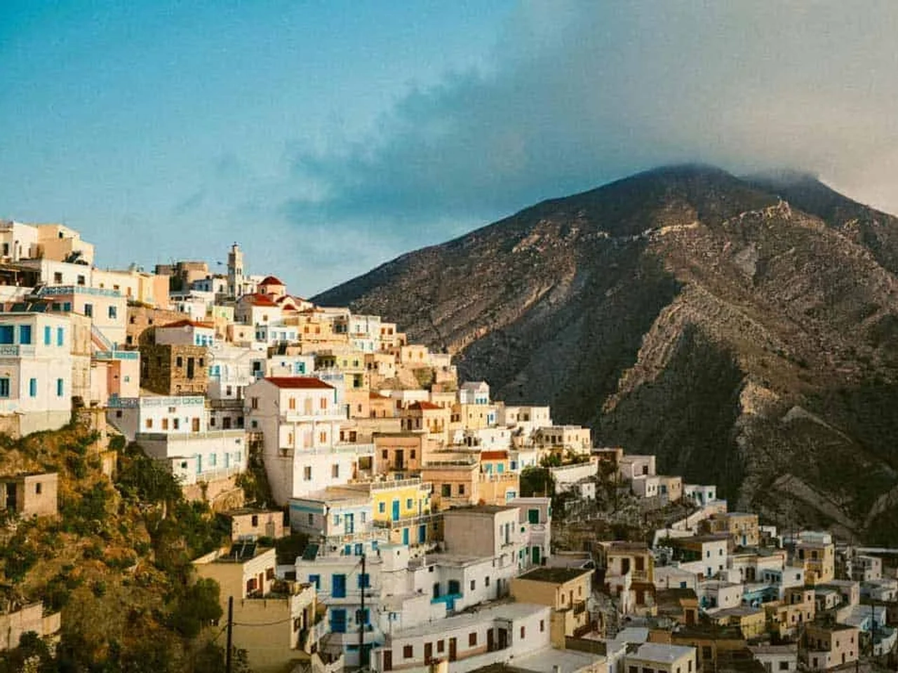 Vista de uma vila grega colorida ao pé de uma montanha. Céu claro ao fundo.