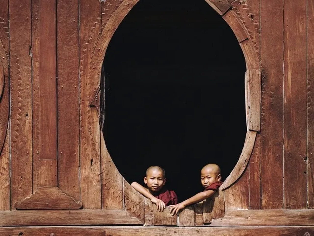 Dos monjes budistas asomándose por una ventana ovalada de madera.