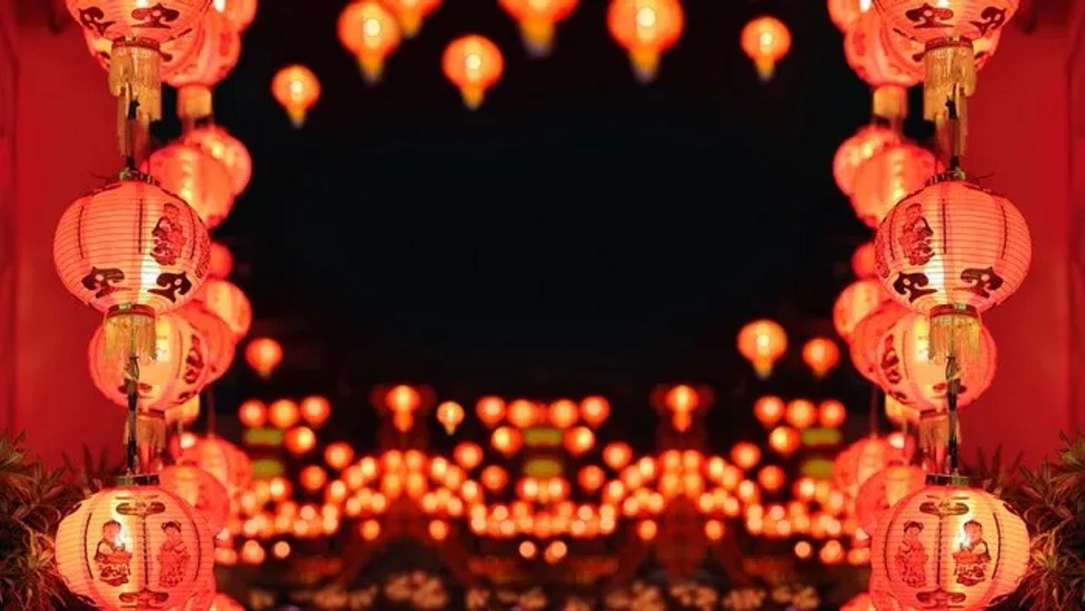 Lanterne rosse cinesi illuminate di notte, appese in file lungo un passaggio, con altre luci sfocate sullo sfondo.