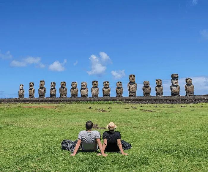 Pareja sentada frente a moáis en la Isla de Pascua bajo un cielo azul.