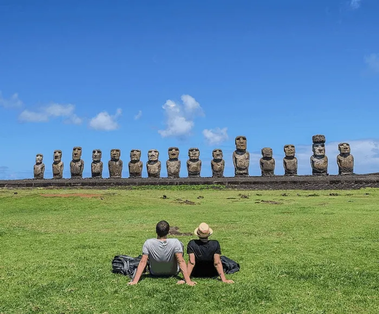 Pareja sentada frente a moáis en la Isla de Pascua bajo un cielo azul.