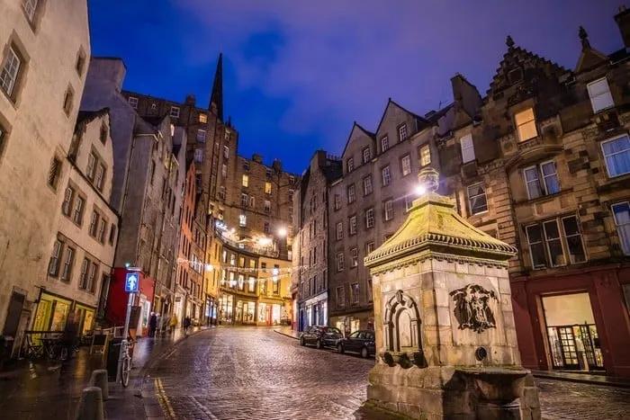 Rua iluminada em Edimburgo, com arquitetura histórica e uma fonte no centro.