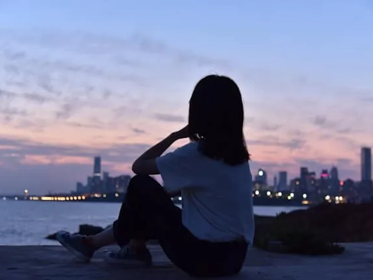 rapariga sentada ao pé do rio com vista para os prédios e o por do sol roxo