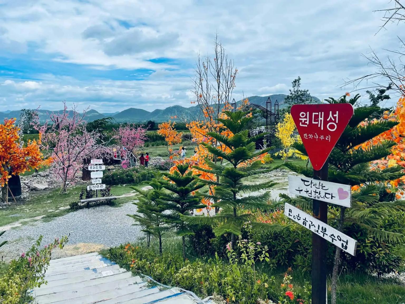 Jardim colorido com flores e sinalização em Kanchanaburi, cercado por montanhas.