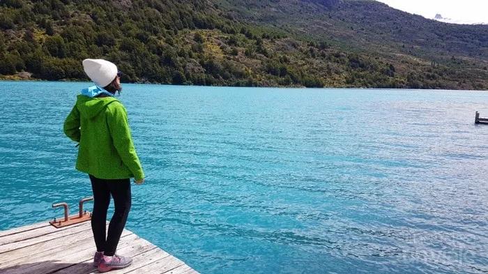 Mujer de pie en un muelle, mirando un lago azul rodeado de montañas.