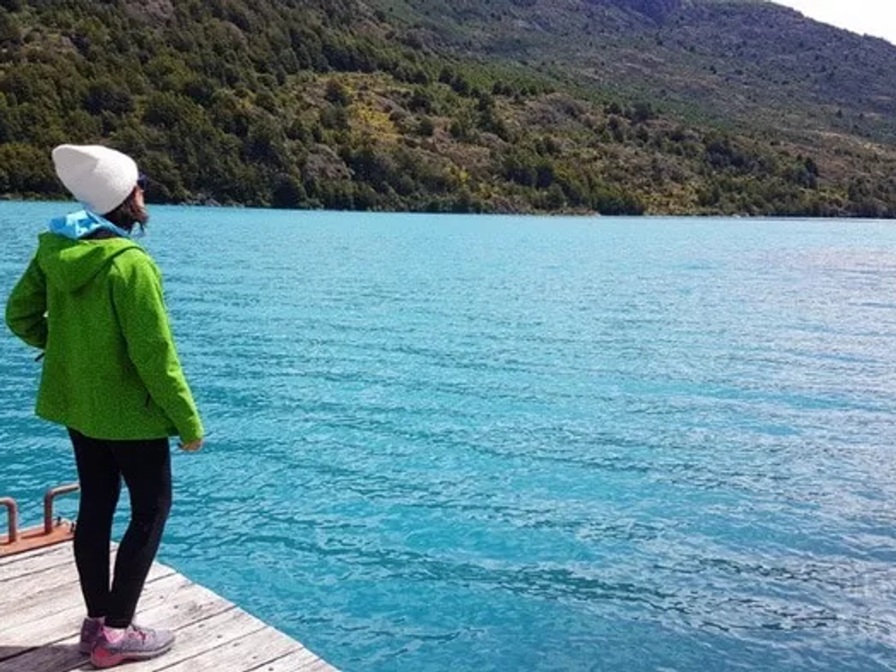 Mujer de pie en un muelle, mirando un lago azul rodeado de montañas.