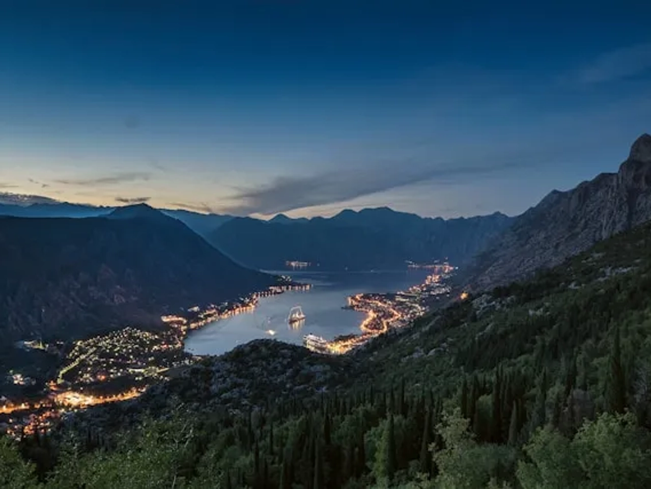 vista de kotor em montenegro ao por do sol, com luzes das casas e barcos a andar no mar