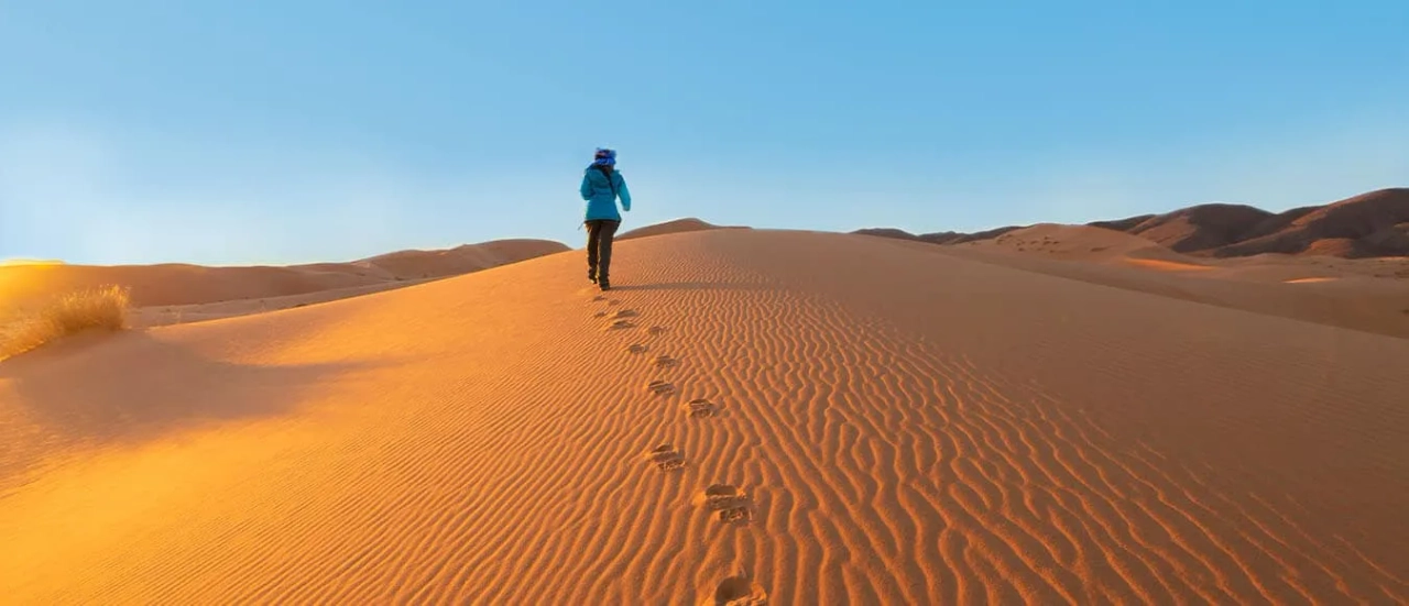 Pessoa caminhando sobre dunas de areia no deserto ao amanhecer.
