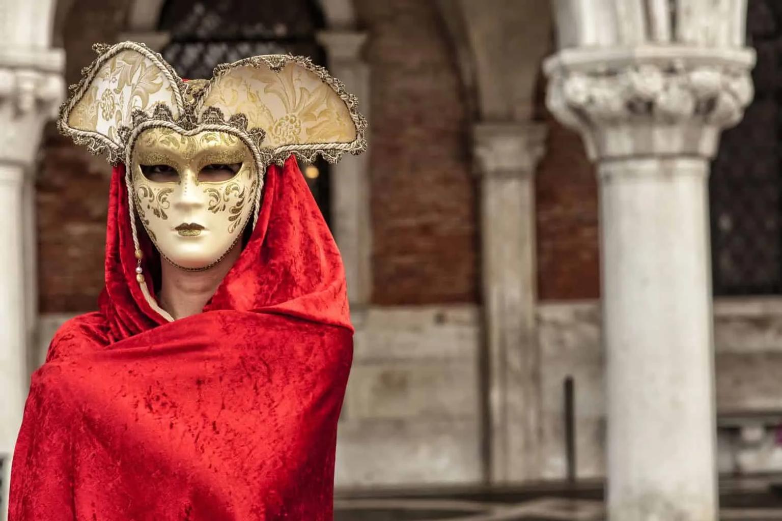 Pessoa com máscara elaborada e manto vermelho, destacando-se em ambiente de Carnaval.