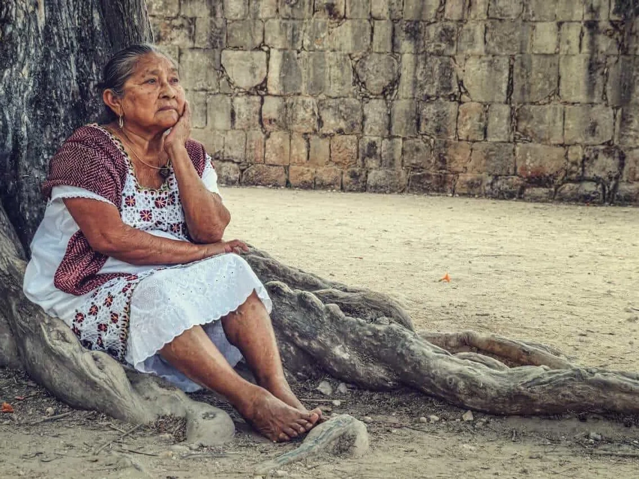 Mulher idosa sentada sob uma árvore, refletindo em um ambiente histórico.