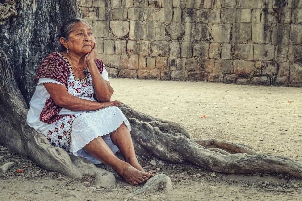 Mulher idosa sentada sob uma árvore, refletindo em um ambiente histórico.