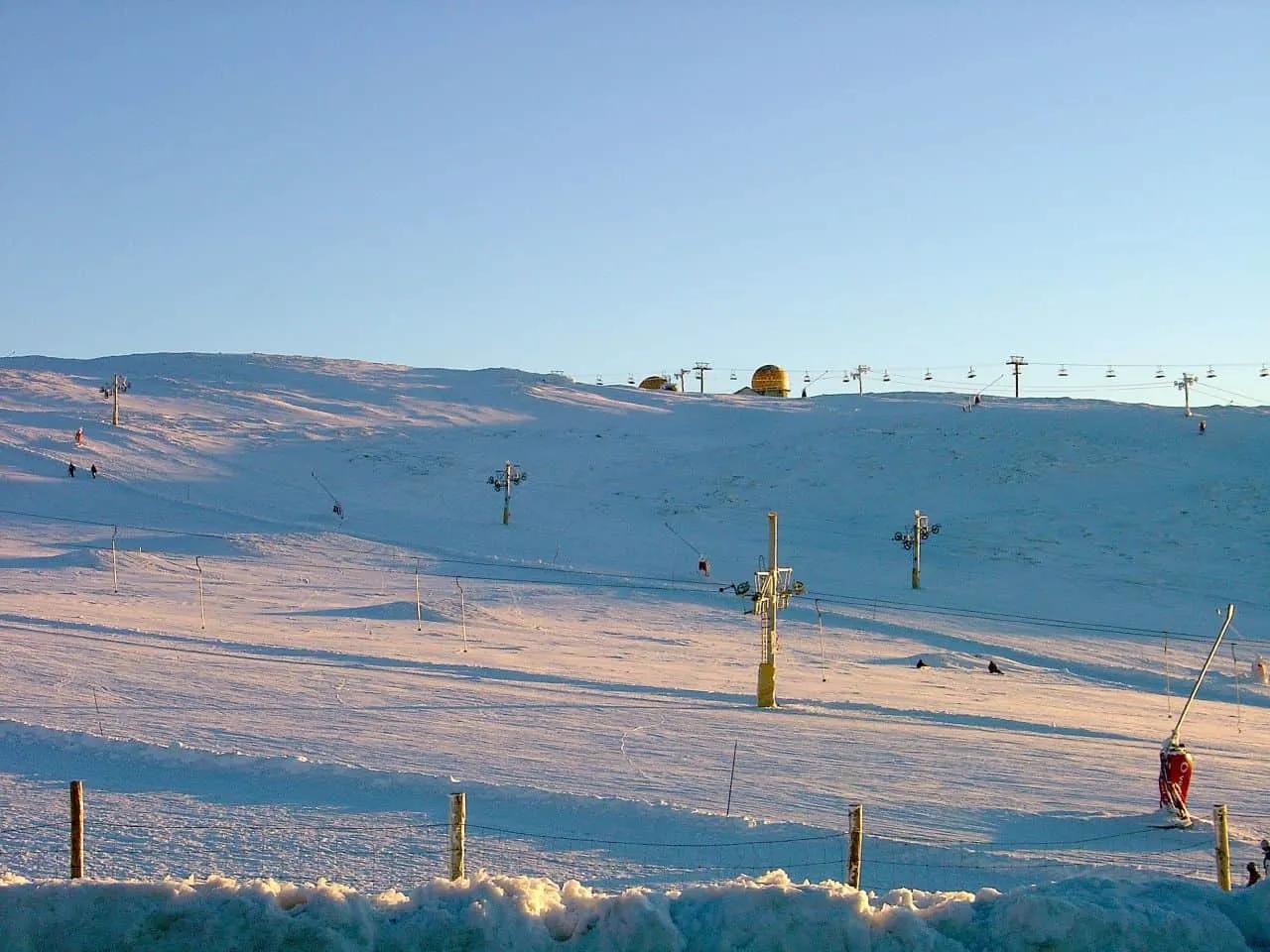 Paisagem de montanha coberta de neve, com esquiadores e teleféricos visíveis.