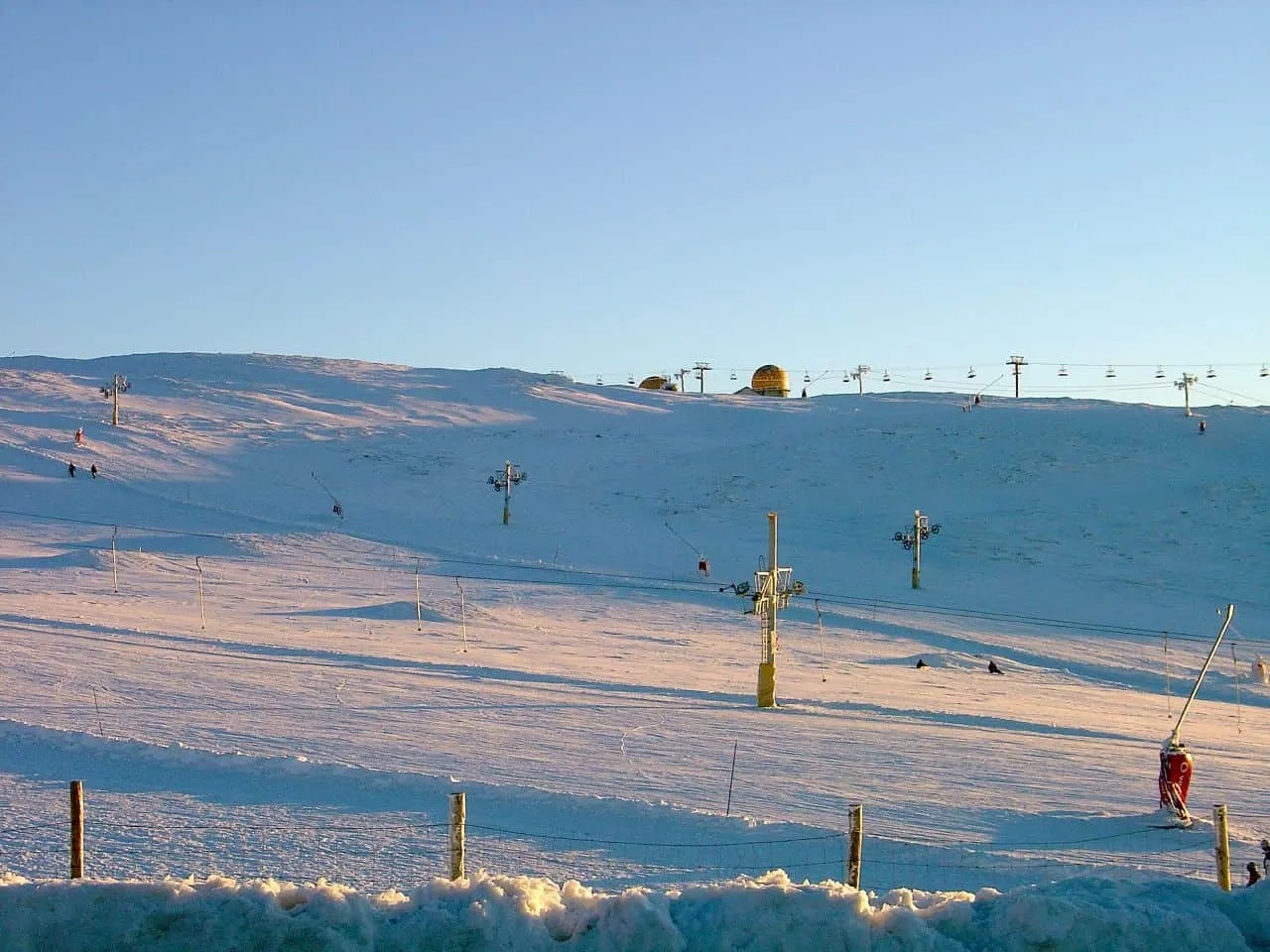 Paisagem de montanha coberta de neve, com esquiadores e teleféricos visíveis.