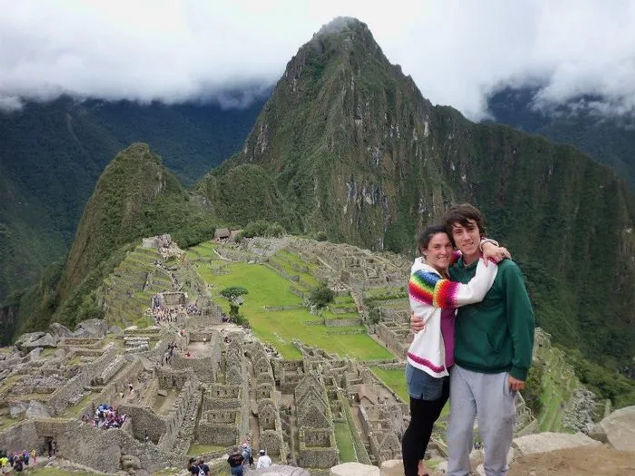 Pareja abrazándose frente a Machu Picchu, con montañas y nubes de fondo.