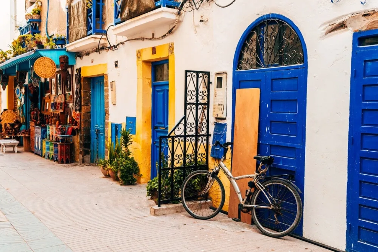 Strada colorata nella medina di Essaouira, con porte blu, facciate bianche, botteghe artigiane e una bicicletta appoggiata al muro.