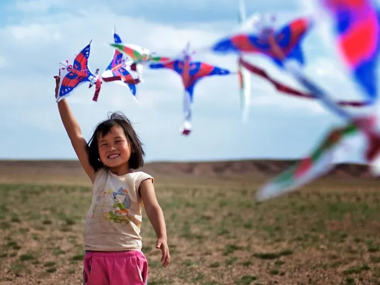 Niña sonriente sosteniendo cometas de colores en un paisaje abierto.