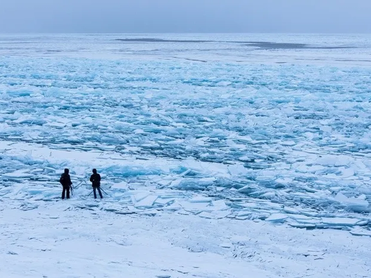 Dos personas observan un lago helado con fragmentos de hielo.