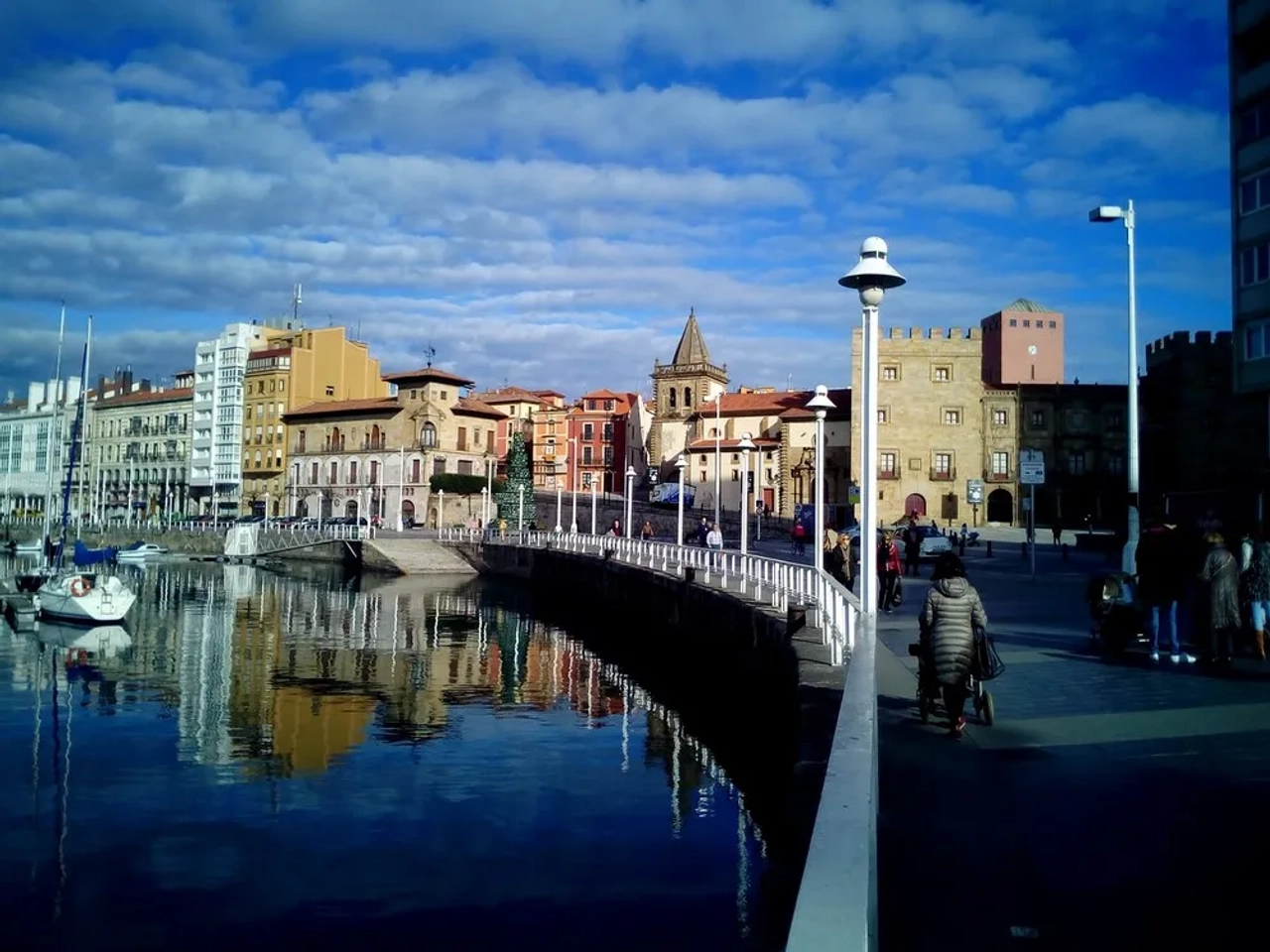 Cais com edifícios coloridos refletidos na água e pessoas caminhando.