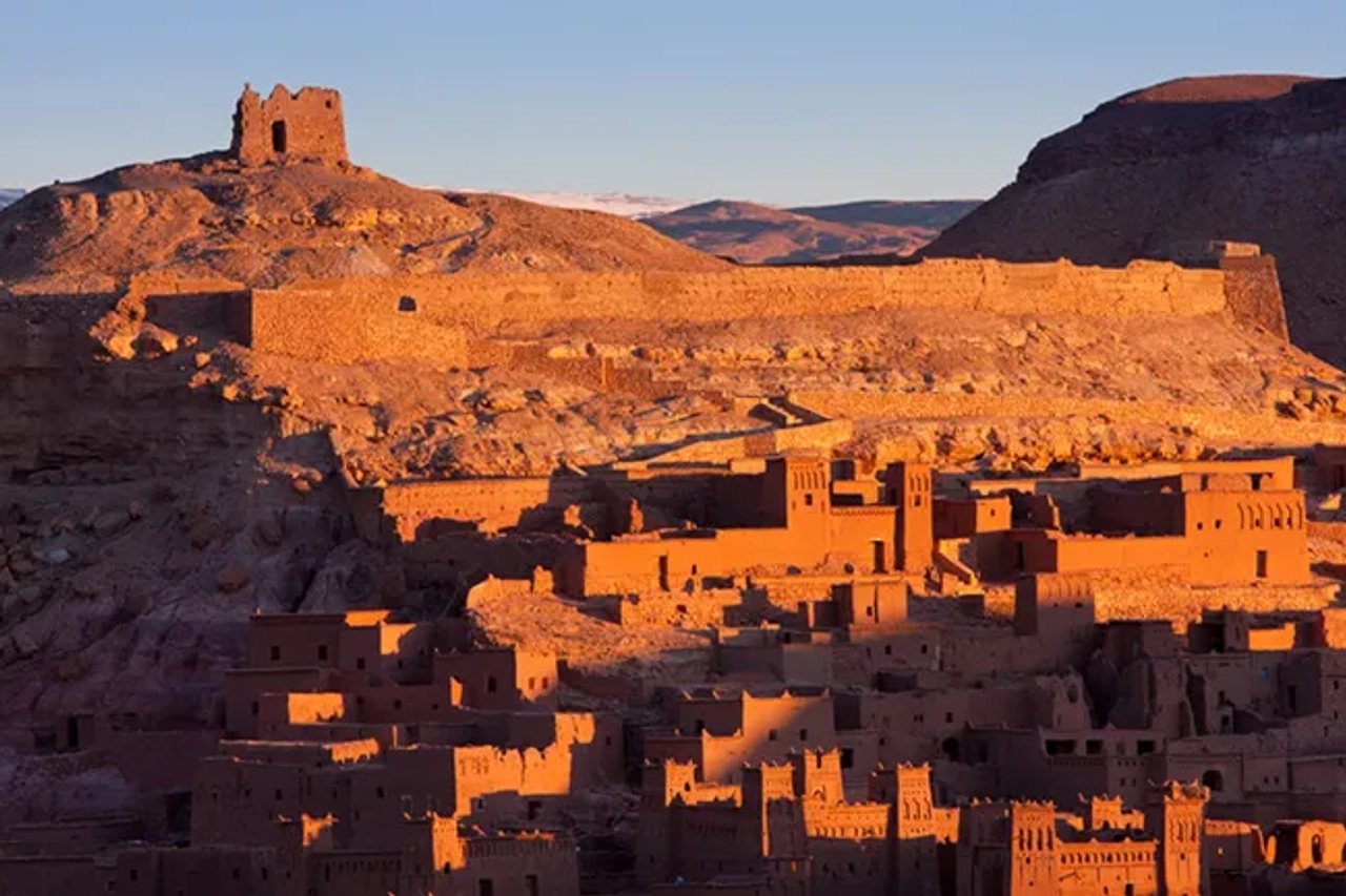 Ksar di Ait Ben Haddou al tramonto, con edifici in terra cruda illuminati dalla luce calda e colline desertiche sullo sfondo.