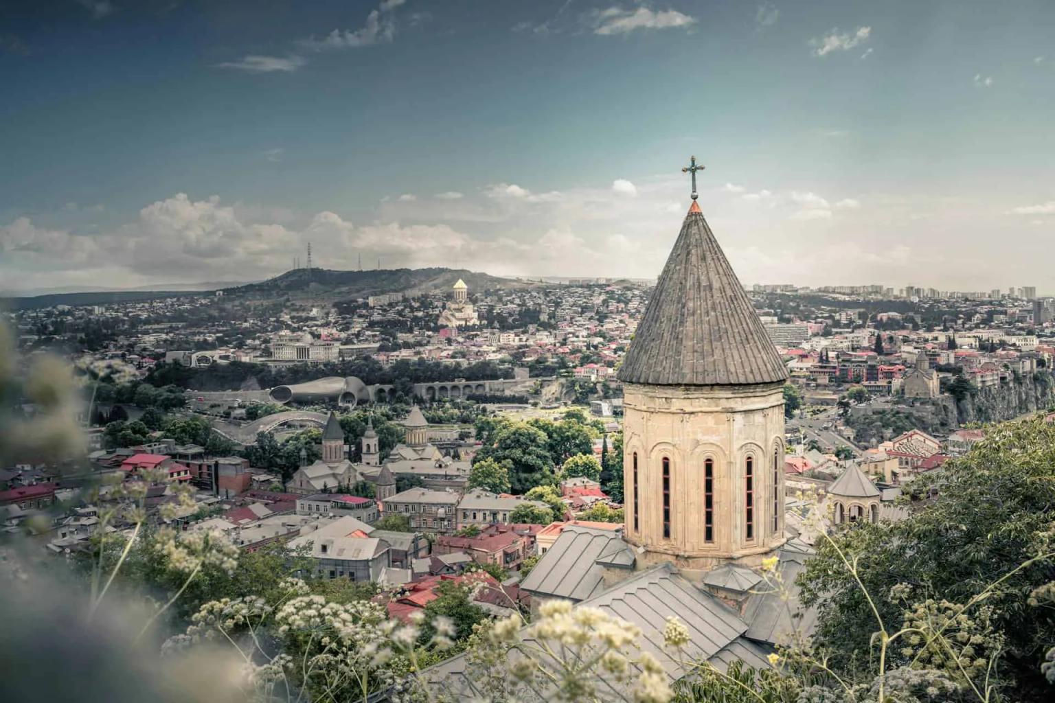 Vista panorâmica de Tbilisi, com destaque para uma igreja e montanhas ao fundo.