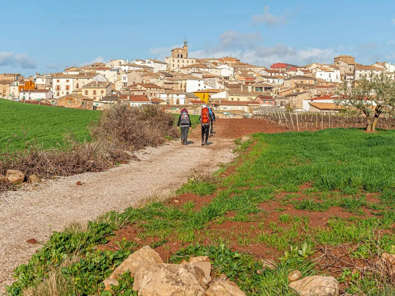 Caminhantes seguem um caminho rural em direção a uma vila. Céu azul ao fundo.