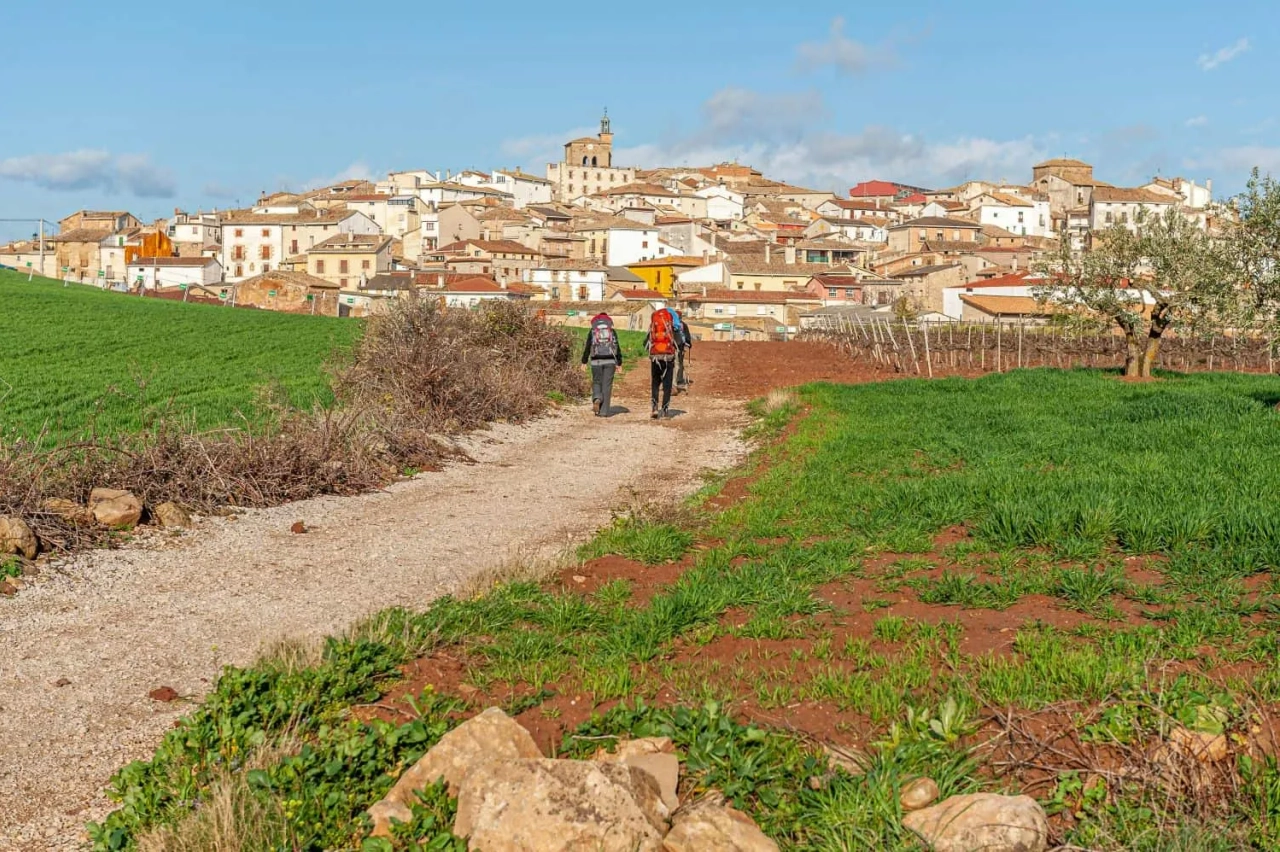 Caminhantes seguem um caminho rural em direção a uma vila. Céu azul ao fundo.