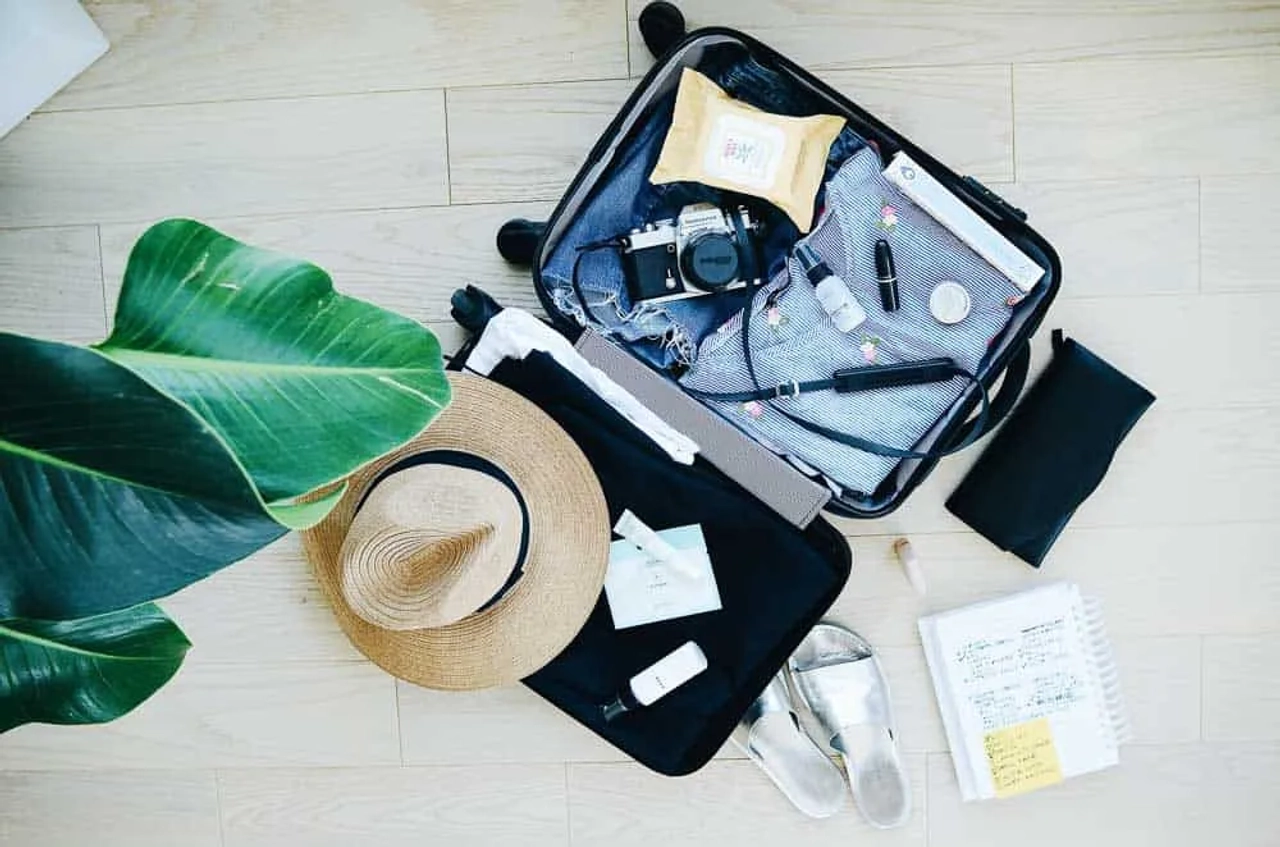 Maleta abierta con ropa, accesorios de viaje y una planta al fondo.