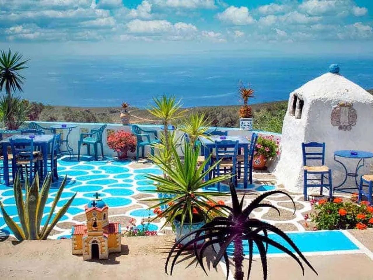 Vista de um terraço grego com mesas, cadeiras e plantas, ao fundo o mar.