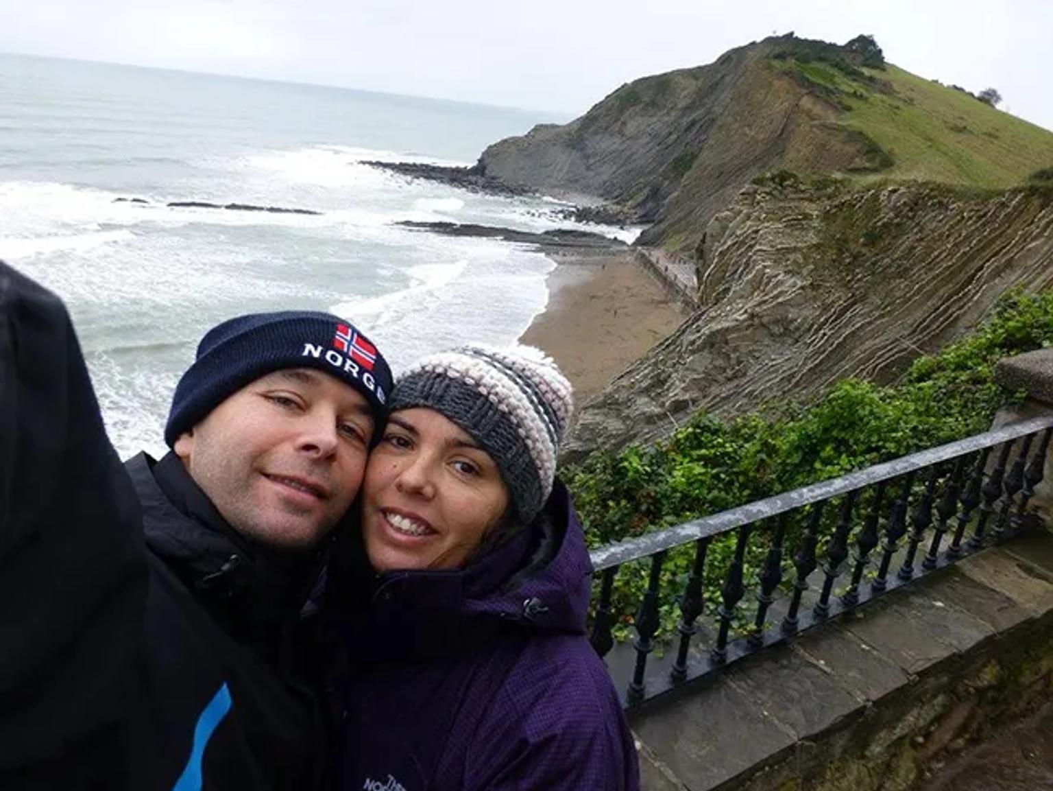 Pareja sonriente en un mirador con vista a la costa y acantilados.